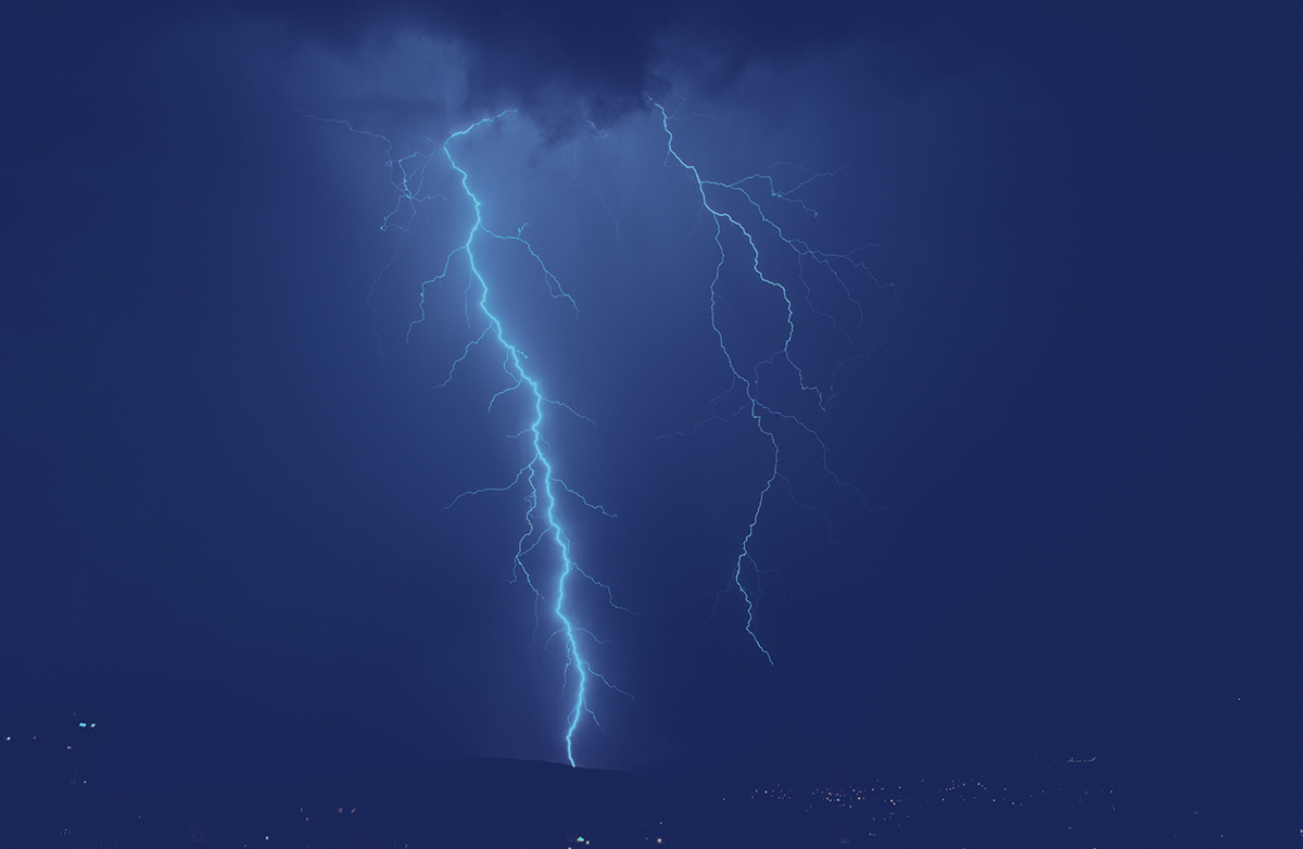 A photograph of a bright blue bolt of lightning striking the ground against a dark night sky. The main bolt features jagged, zigzagging branches that split into thinner and thinner electrical paths as they move away from the center, showcasing a natural fractal structure.