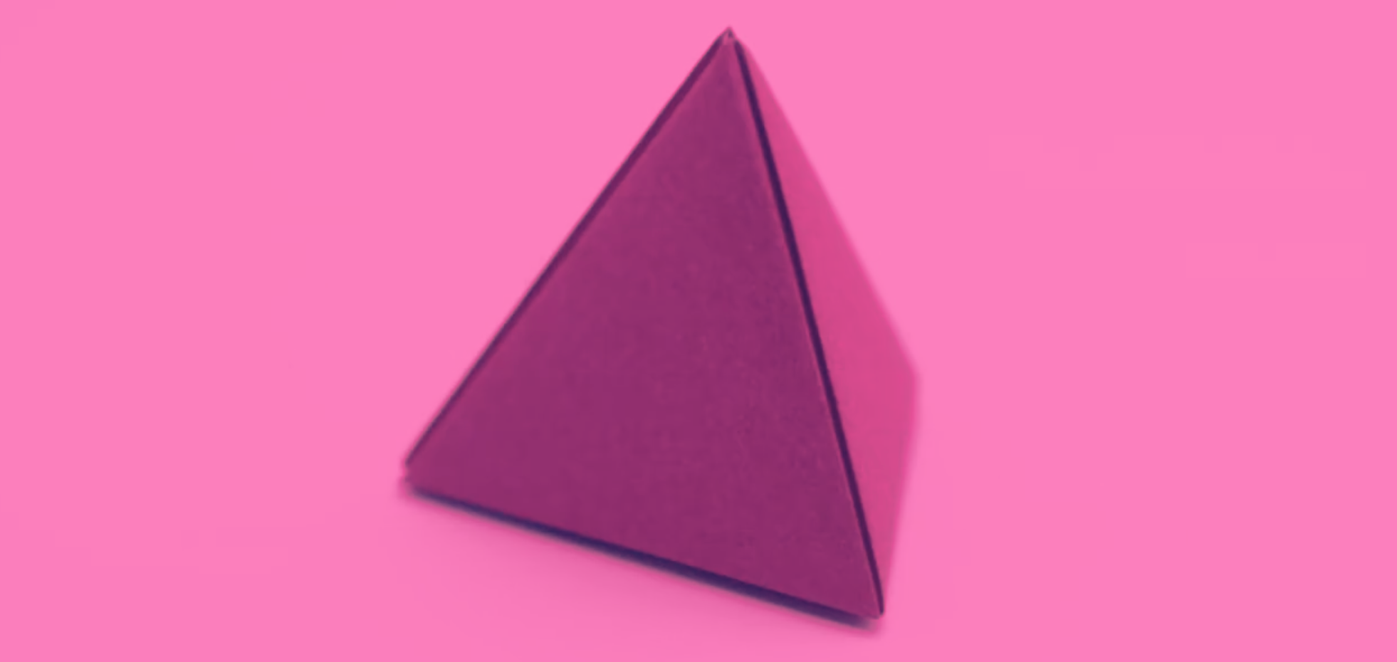 A close-up photograph of a three-dimensional tetrahedron made from pink paper, set against a matching pink background. The shape is a triangular pyramid with four equal triangular faces, used here as a physical model for geometric exploration.