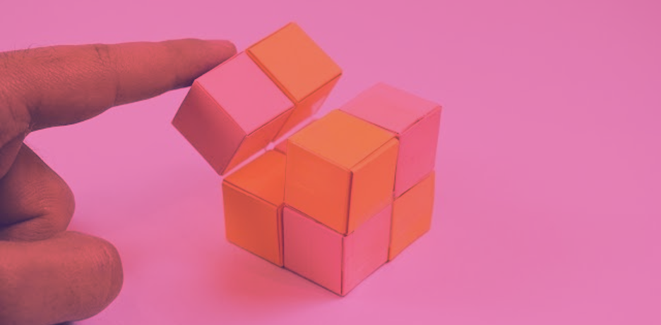 A close-up photograph featuring a hand interacting with several small, colorful paper cubes on a pink surface. The cubes are a mix of bright orange and pink. One cube is being tilted or lifted by a finger, showing how the individual geometric units can be moved or arranged.