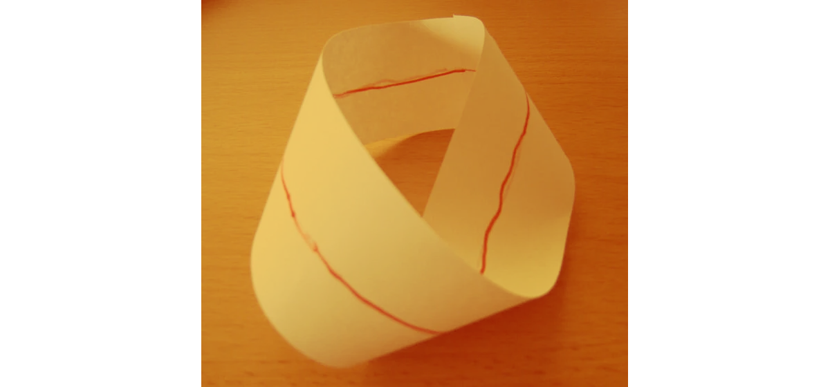 Una fotografía de una banda de Möbius de papel blanco sobre una superficie de madera. Se ha dibujado una fina línea roja a lo largo del centro de todo el bucle, siguiendo su trayectoria de una sola cara para demostrar que la línea finalmente se encuentra con su punto de partida sin cruzar nunca un borde.